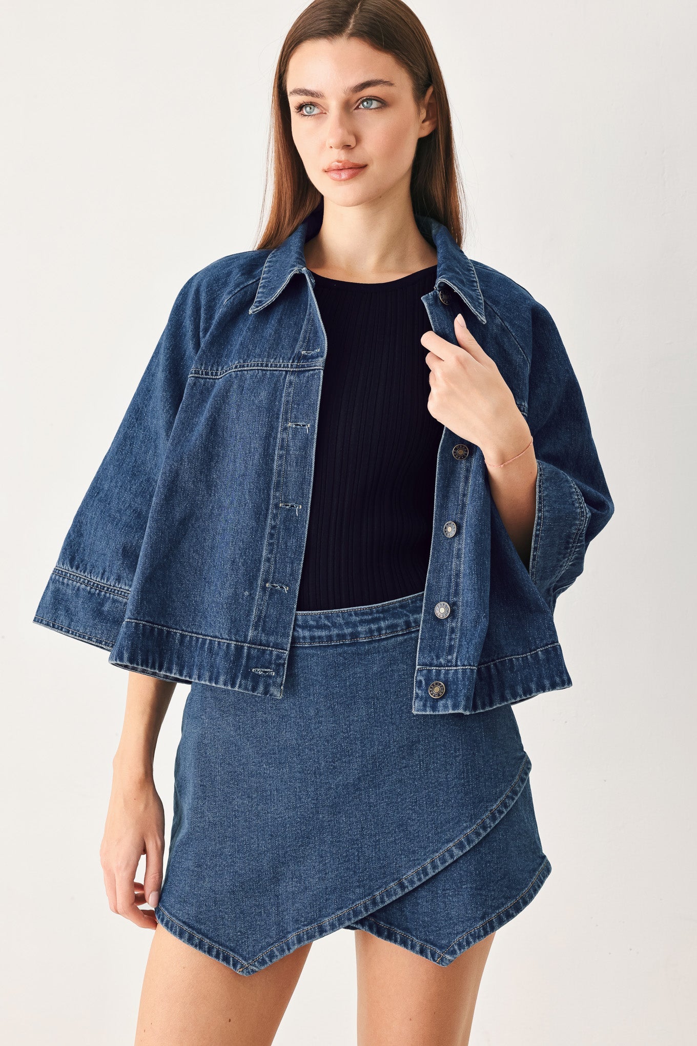 A woman modeling a dark blue cropped denim jacket with wide sleeves, paired with a matching denim mini skirt and a black ribbed top, against a plain light background.