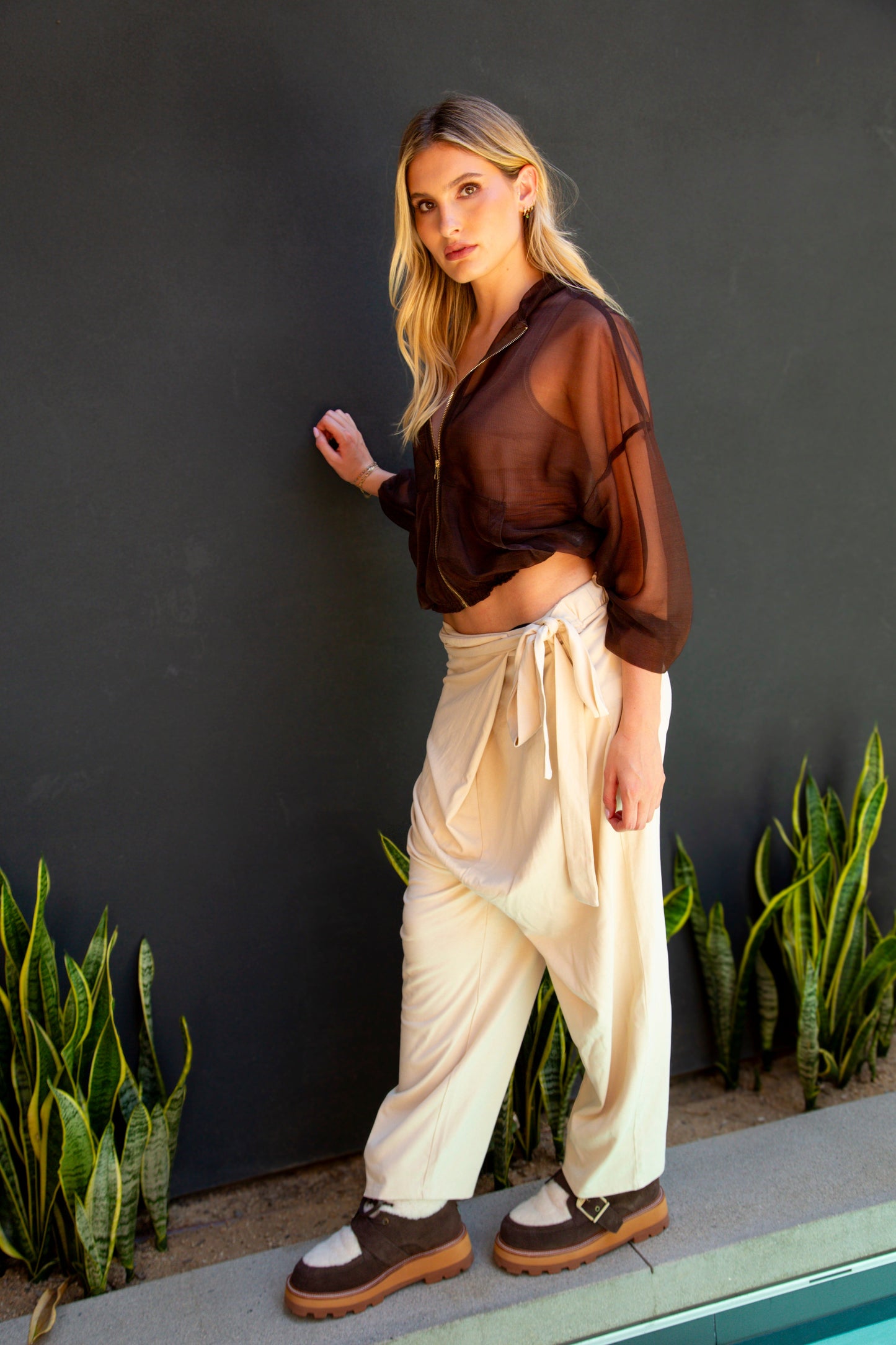 Woman in a sheer brown top and white pants standing against a dark wall with plants.