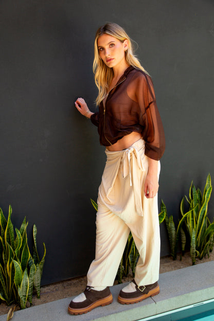 Woman in a sheer brown top and white pants standing against a dark wall with plants.
