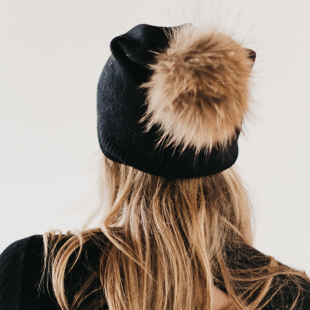 Black beanie with a fur pom-pom worn by a person with long blonde hair on a white background