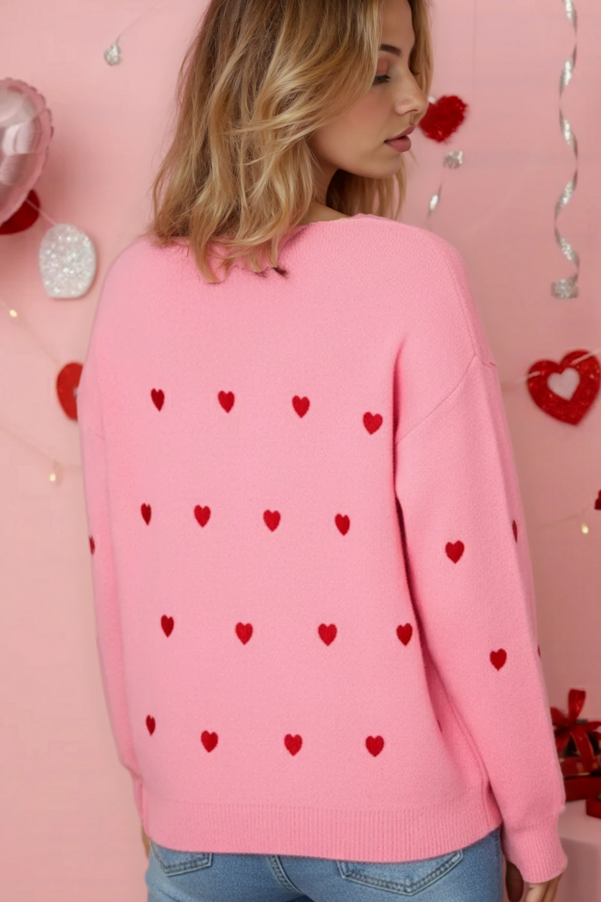 Person wearing a pink sweater with red heart patterns against a pink background with heart decorations.