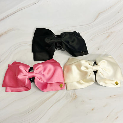 Three large satin hair bows in black, pink, and white on a light wooden surface.