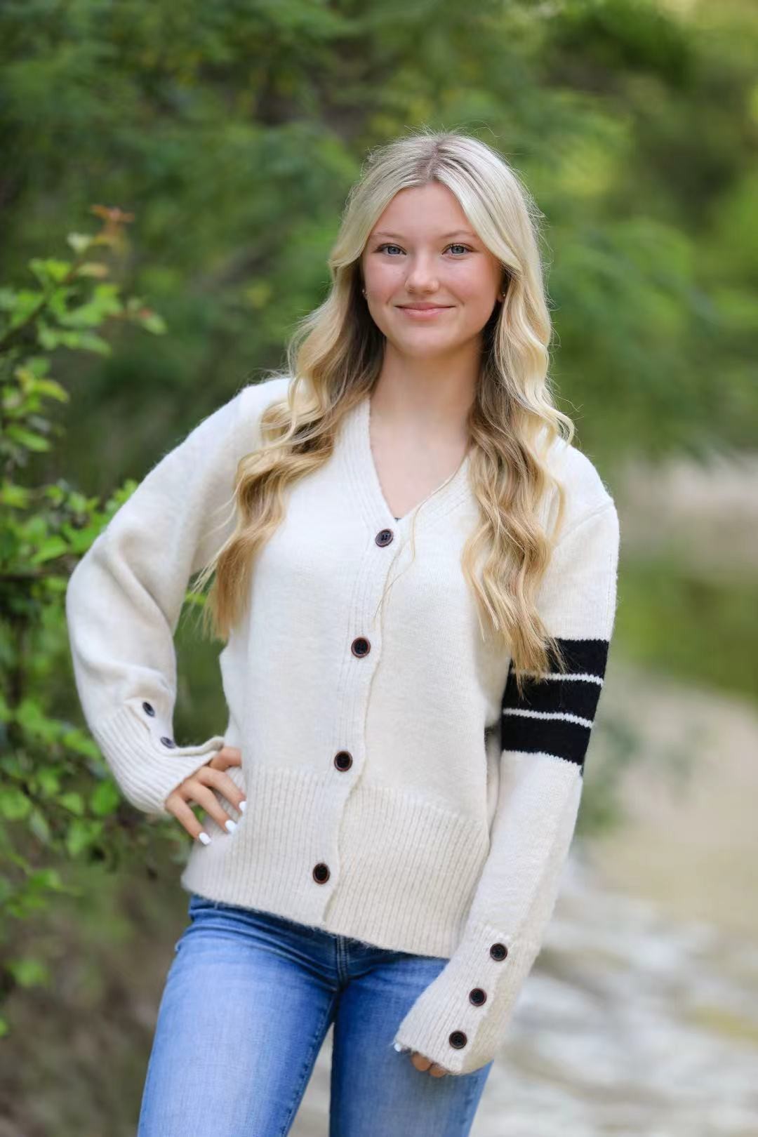 Woman wearing a white cardigan with black buttons and sleeves, standing outdoors with greenery in the background.