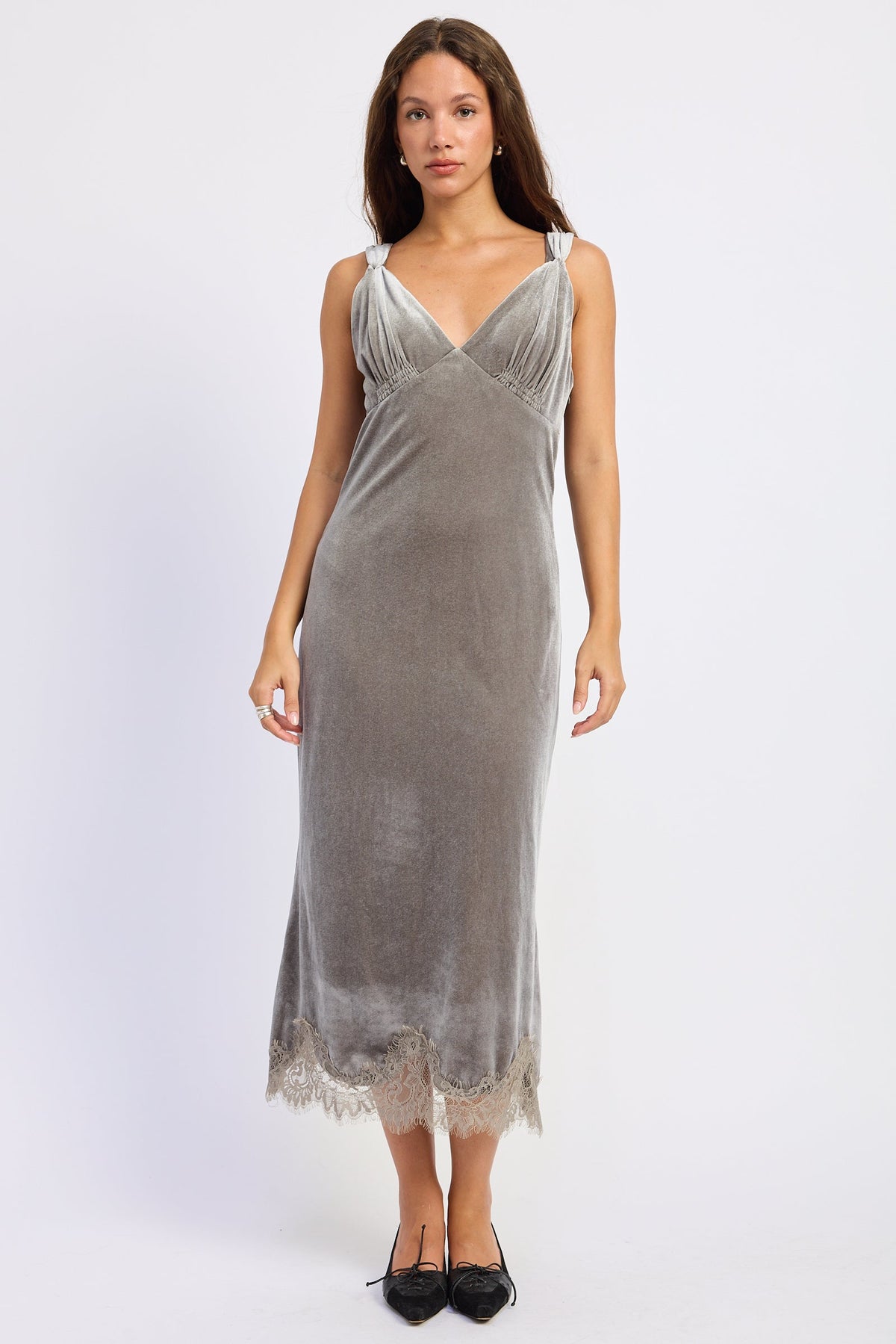 A woman modeling a sleeveless gray velvet slip dress with a V-neckline, gathered bodice, and lace-trimmed hem, paired with black pointed flats against a light gray studio background.