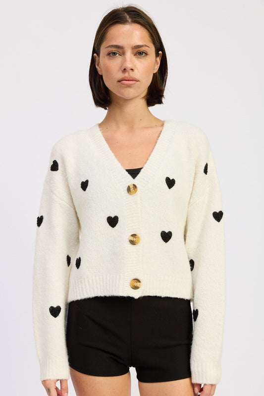 Woman wearing a cream cardigan with black heart patterns and gold buttons on a light gray background