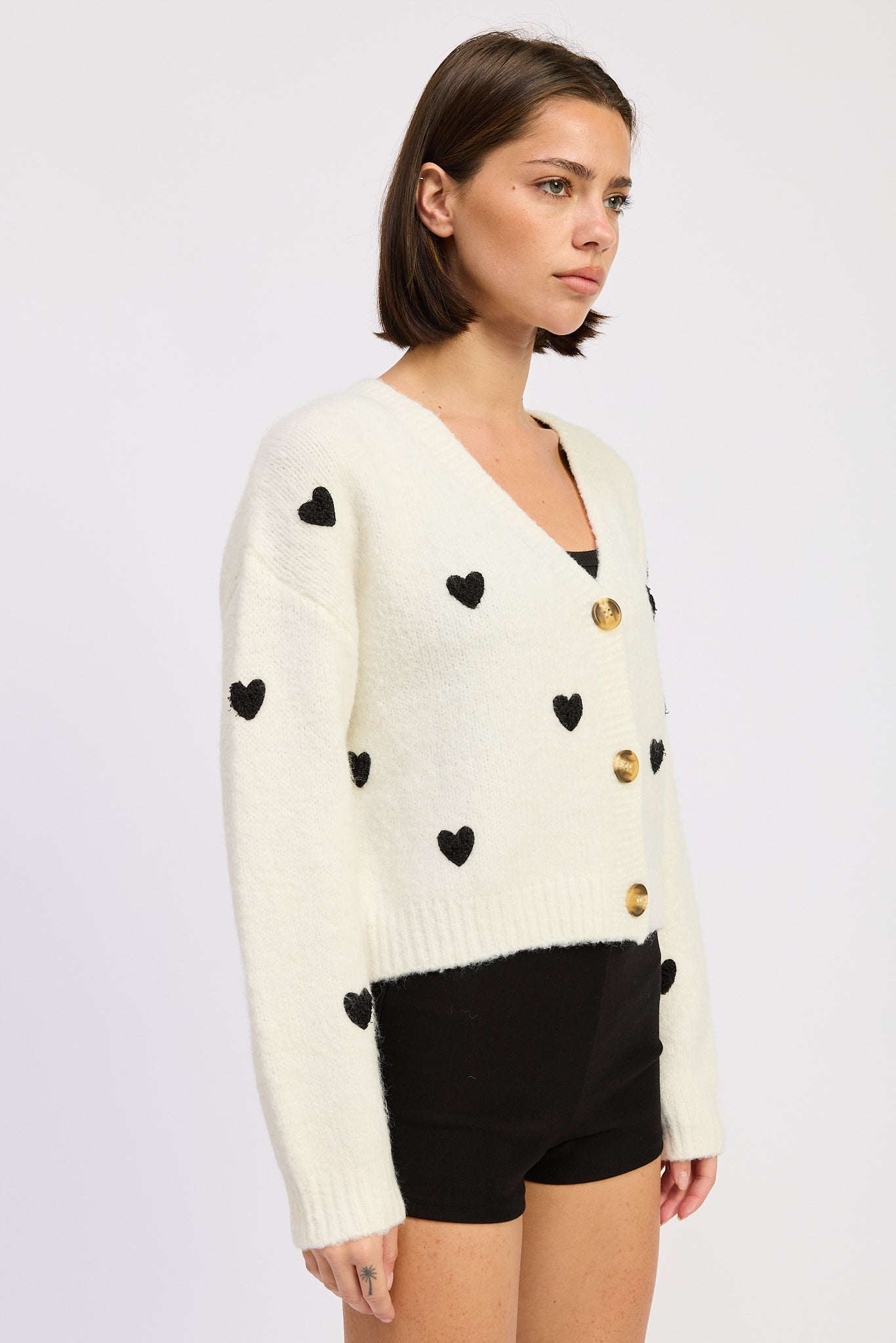 Woman wearing a cream sweater with black heart patterns and gold accents on a white background