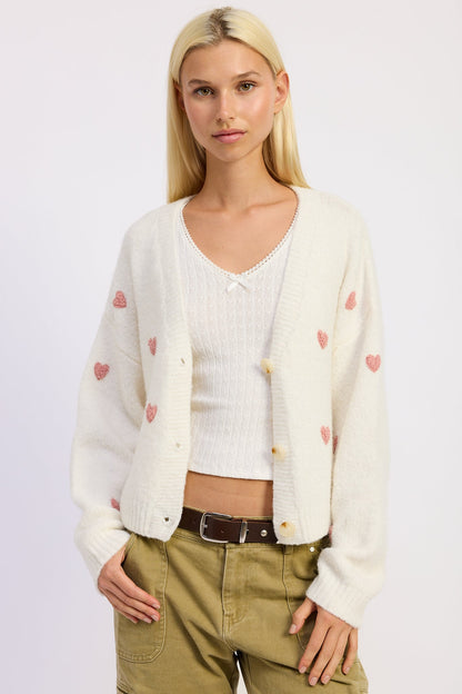 Woman wearing a cream cardigan with heart patterns over a white shirt on a light background