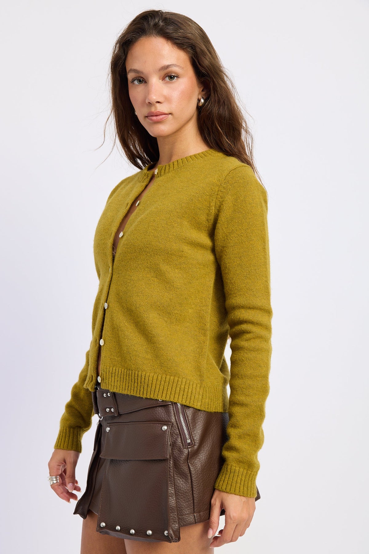 A woman modeling a mustard yellow knit cardigan sweater with brown buttons, paired with a brown mini skirt and a matching brown crossbody bag, against a plain light gray studio background.