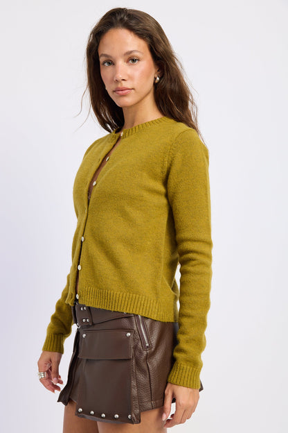 A woman modeling a mustard yellow knit cardigan sweater with brown buttons, paired with a brown mini skirt and a matching brown crossbody bag, against a plain light gray studio background.