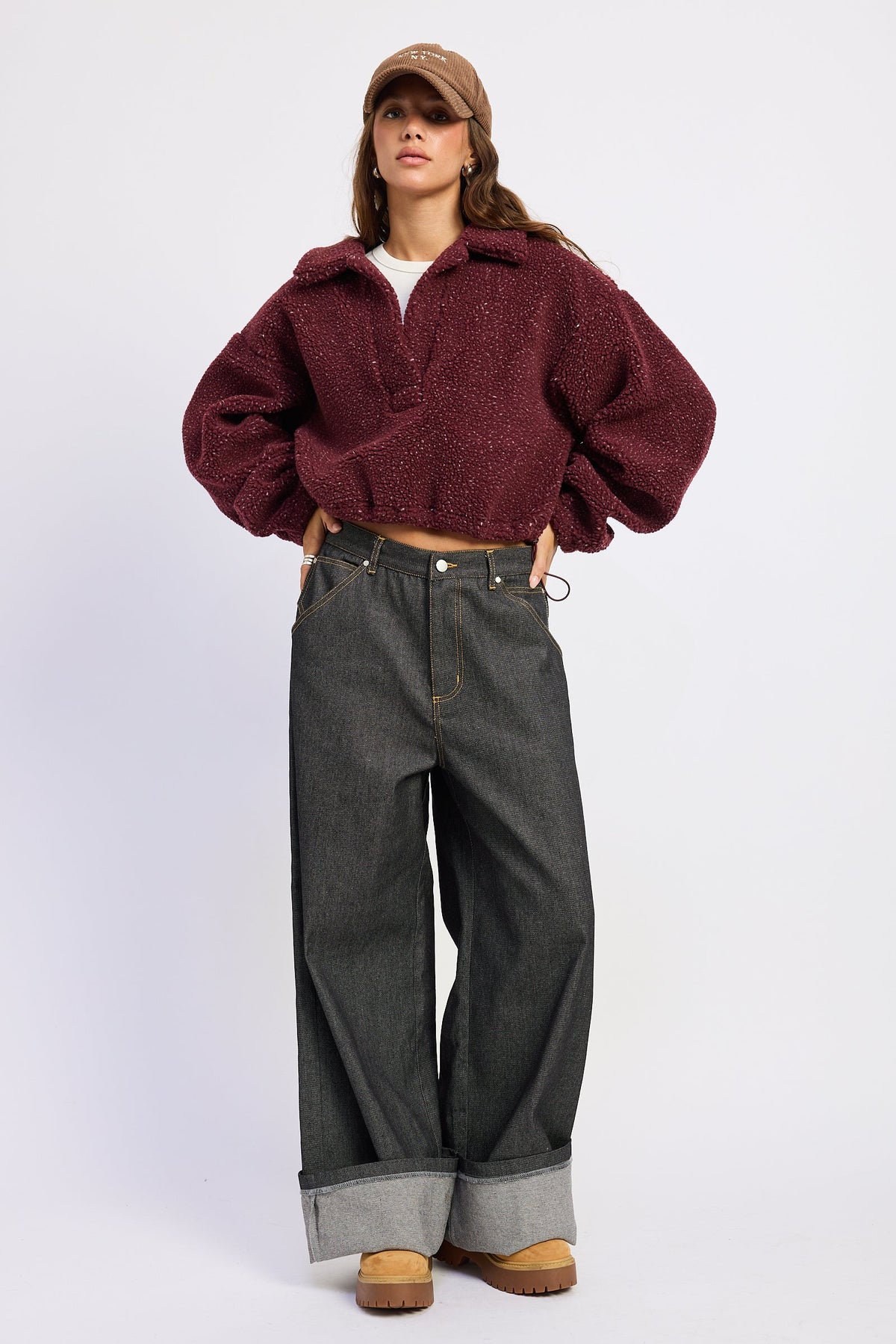 A model wearing a dark burgundy fuzzy cropped jacket, dark gray wide-leg jeans, a brown cap, and brown boots, standing against a light gray studio background.