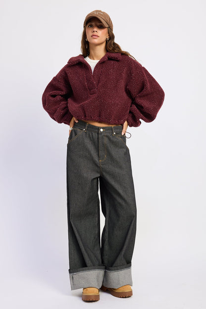 A model wearing a dark burgundy fuzzy cropped jacket, dark gray wide-leg jeans, a brown cap, and brown boots, standing against a light gray studio background.