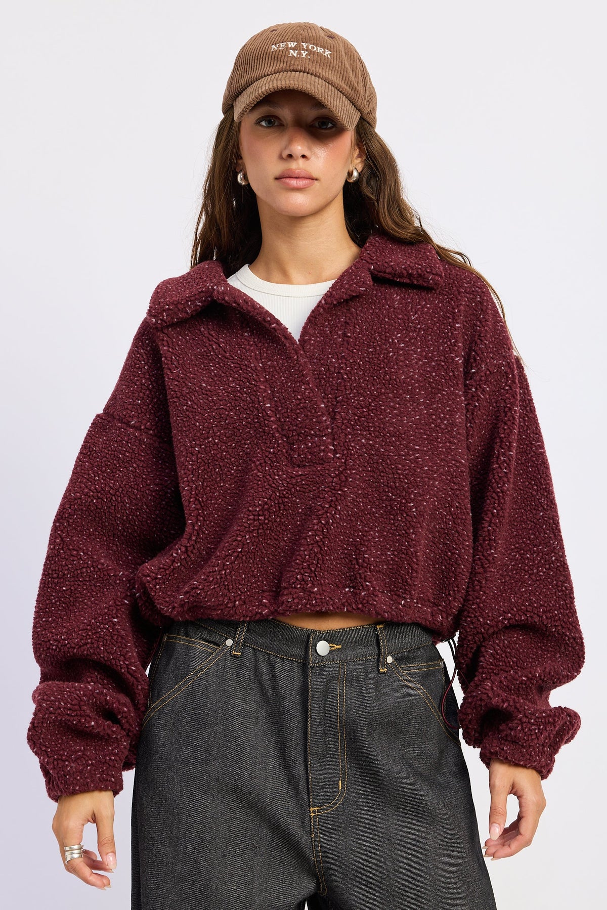 A woman wearing a textured, cropped maroon fleece zip-up jacket, a brown embroidered baseball cap, and dark gray jeans, photographed against a light gray studio background.