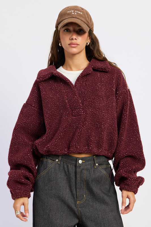 A woman wearing a textured, cropped maroon fleece zip-up jacket, a brown embroidered baseball cap, and dark gray jeans, photographed against a light gray studio background.