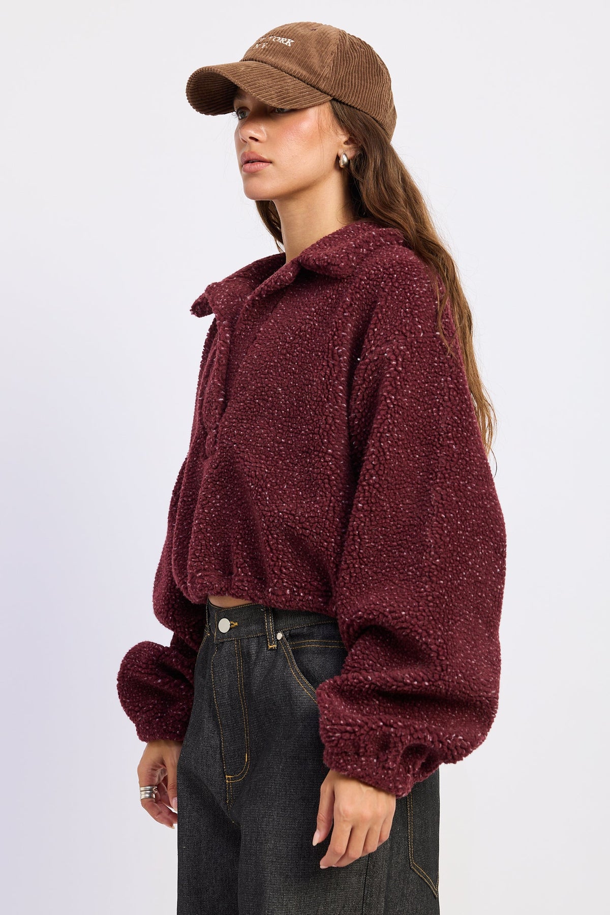 A woman wearing a textured, cropped maroon fleece pullover, dark gray jeans, and a brown corduroy cap, shown from a side profile against a light gray studio background.