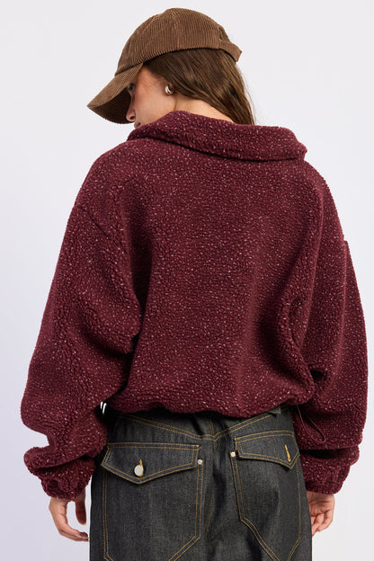 A person shown from the back wearing a burgundy sherpa fleece cropped hoodie, dark blue jeans, and a brown corduroy cap against a light gray studio background.