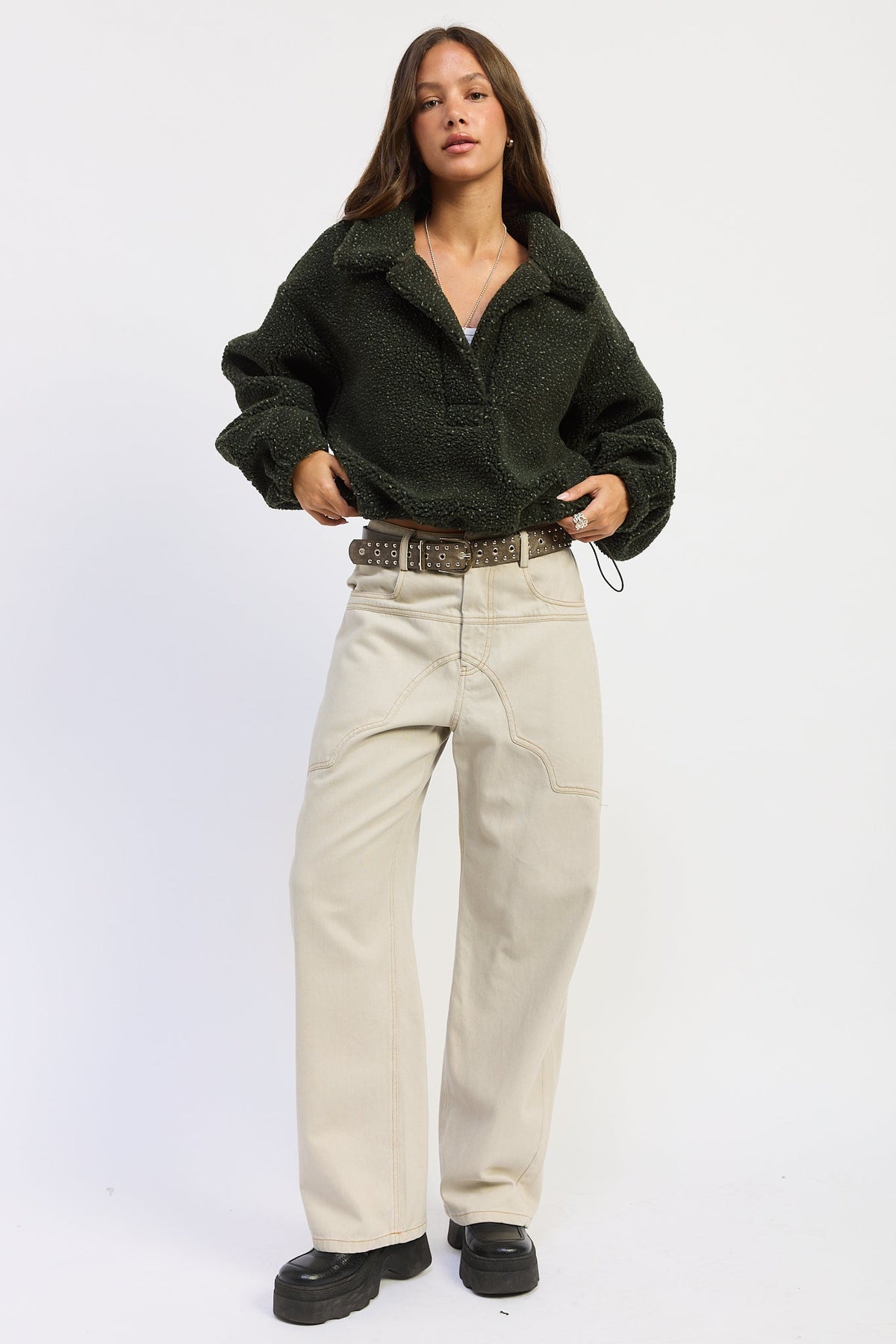 A woman modeling a dark olive green sherpa fleece button-up shirt, paired with beige wide-leg jeans, a brown patterned belt, and black platform shoes, against a plain white studio background.