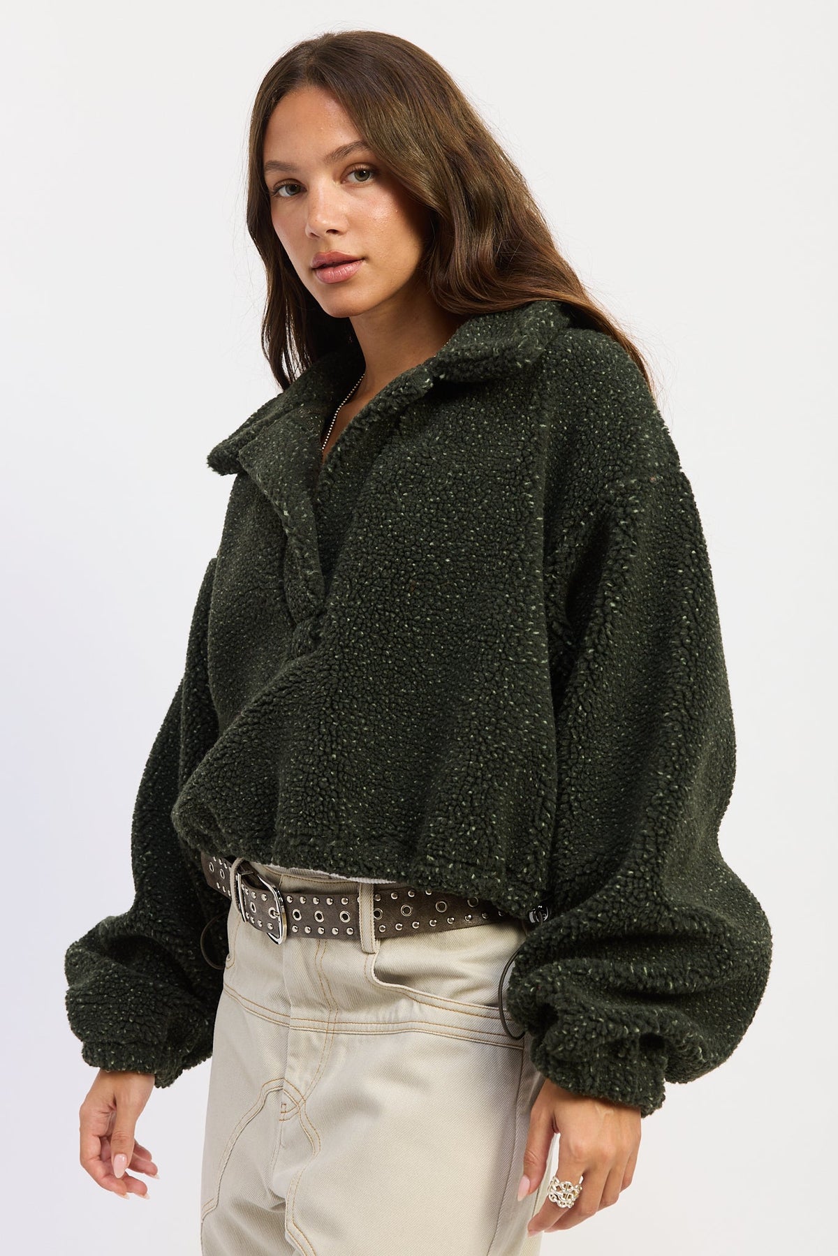 A woman wearing a dark olive green teddy fleece pullover jacket with a half-zip collar, paired with light beige pants and a studded belt, against a plain white studio background.
