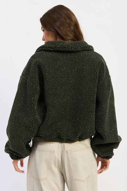 A woman shown from the back wearing a dark olive green textured fleece cropped jacket and cream-colored pants, set against a plain light gray studio background.