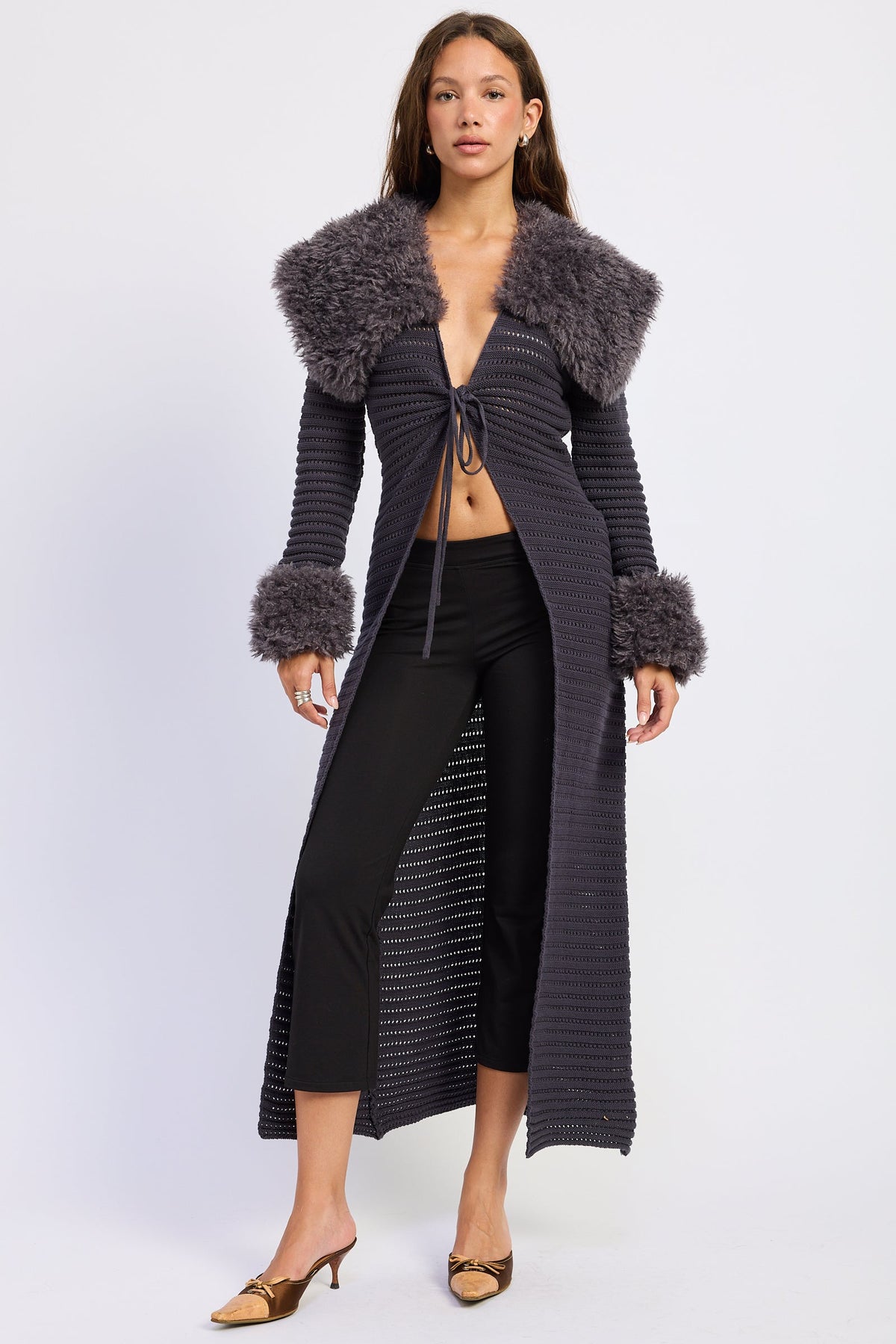 A woman models a long, dark gray ribbed knit cardigan with a thick gray faux fur collar and matching faux fur cuffs, paired with black pants and brown heels against a light gray studio background.