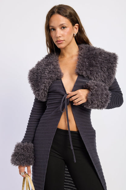 A woman models a dark gray ribbed knit cardigan featuring a large shaggy faux-fur collar and cuffs, styled with black pants against a light gray studio background.