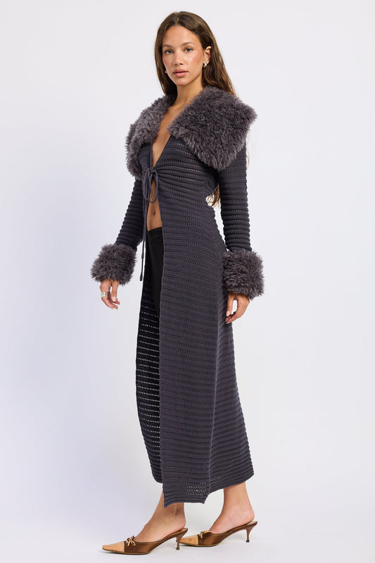 A model wears a long, dark gray ribbed knit wrap coat featuring a voluminous dark gray faux fur collar and matching faux fur cuffs, styled over a black dress and gold mule heels against a light gray studio background.