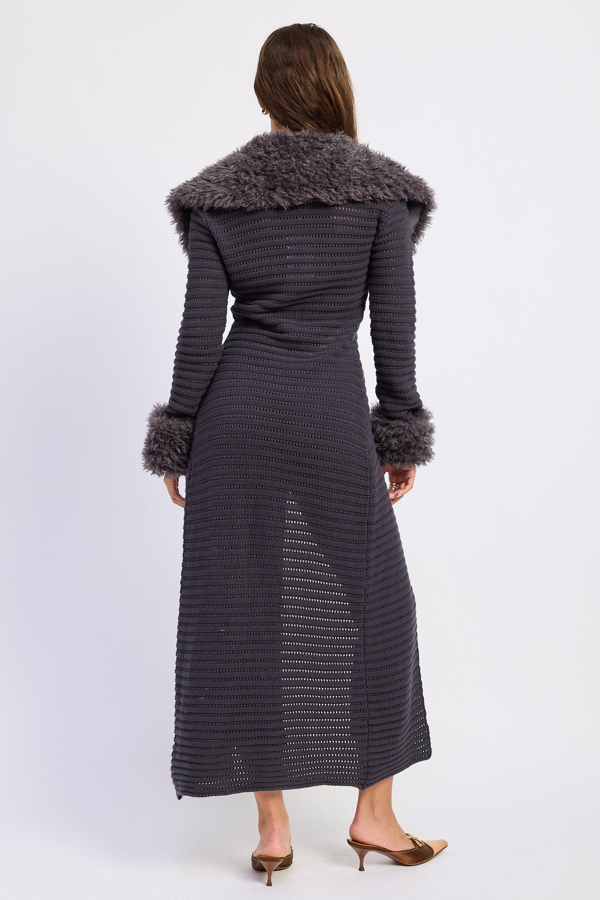 A woman viewed from the back wearing a long dark charcoal ribbed knit dress with a plush gray faux-fur collar and matching faux-fur cuffs, paired with brown pointed-toe heels against a light gray studio background.