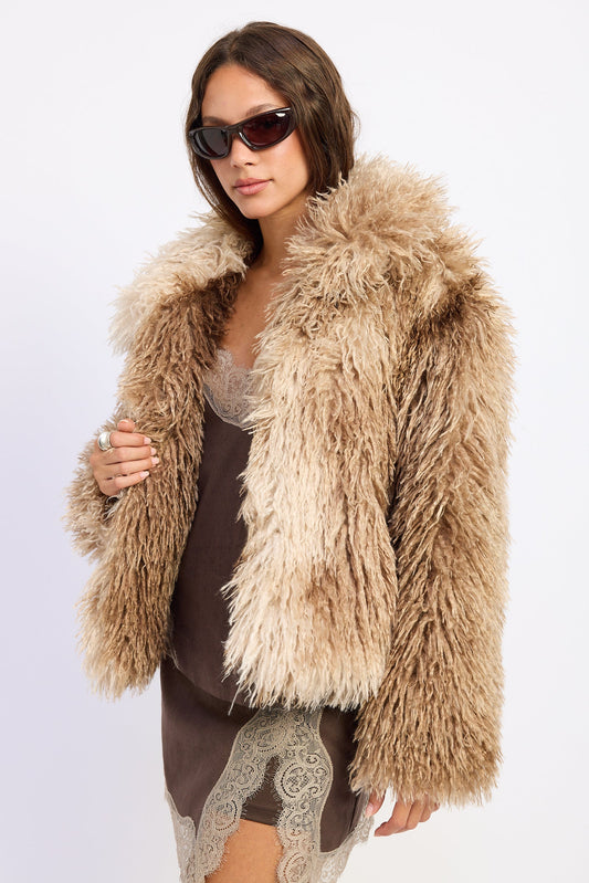 A woman modeling a shaggy, long-pile faux fur jacket in a light tan/beige color, worn over a brown dress with lace trim, set against a plain white studio background.
