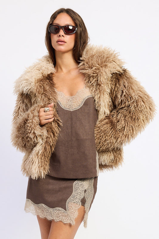 A woman modeling a brown faux-suede slip dress with beige lace trim, styled with a voluminous beige shaggy faux-fur jacket and dark sunglasses against a plain white background.