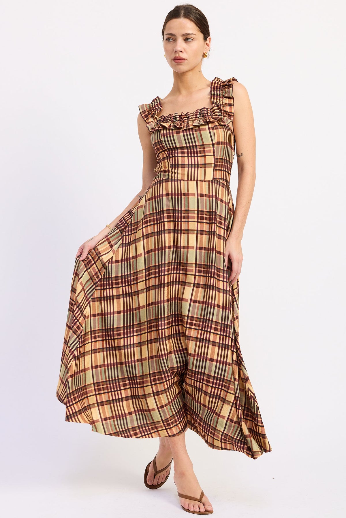 A woman modeling a sleeveless midi dress with a ruffled neckline and a brown, beige, and green plaid pattern, paired with brown flat sandals against a plain white studio background.
