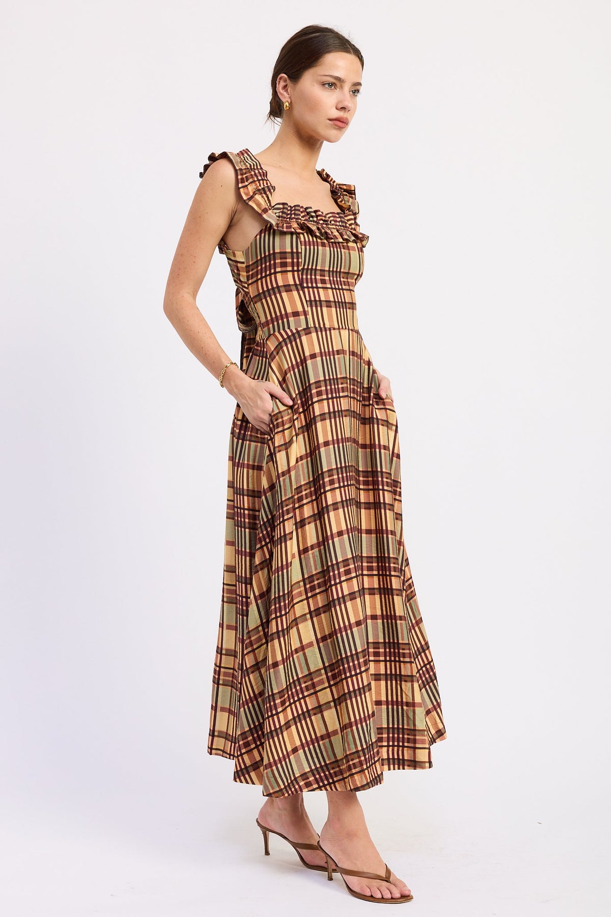 A woman modeling a sleeveless plaid maxi dress with ruffled shoulder straps and side pockets, paired with brown heeled sandals, against a plain white studio background.