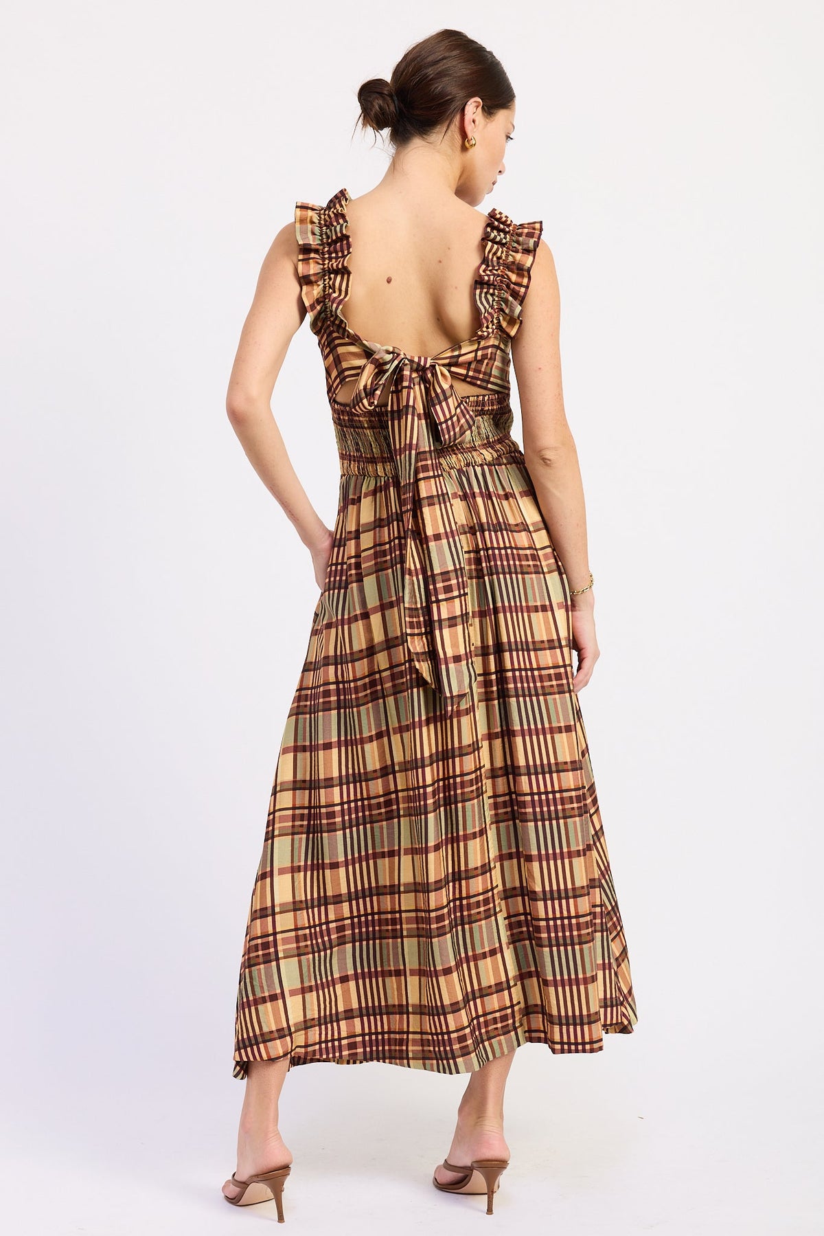 A woman shown from the back wearing a sleeveless maxi dress with ruffled shoulder straps, a tie detail at the back neckline, and a brown, beige, and red plaid pattern, paired with brown heeled sandals against a plain white studio background.