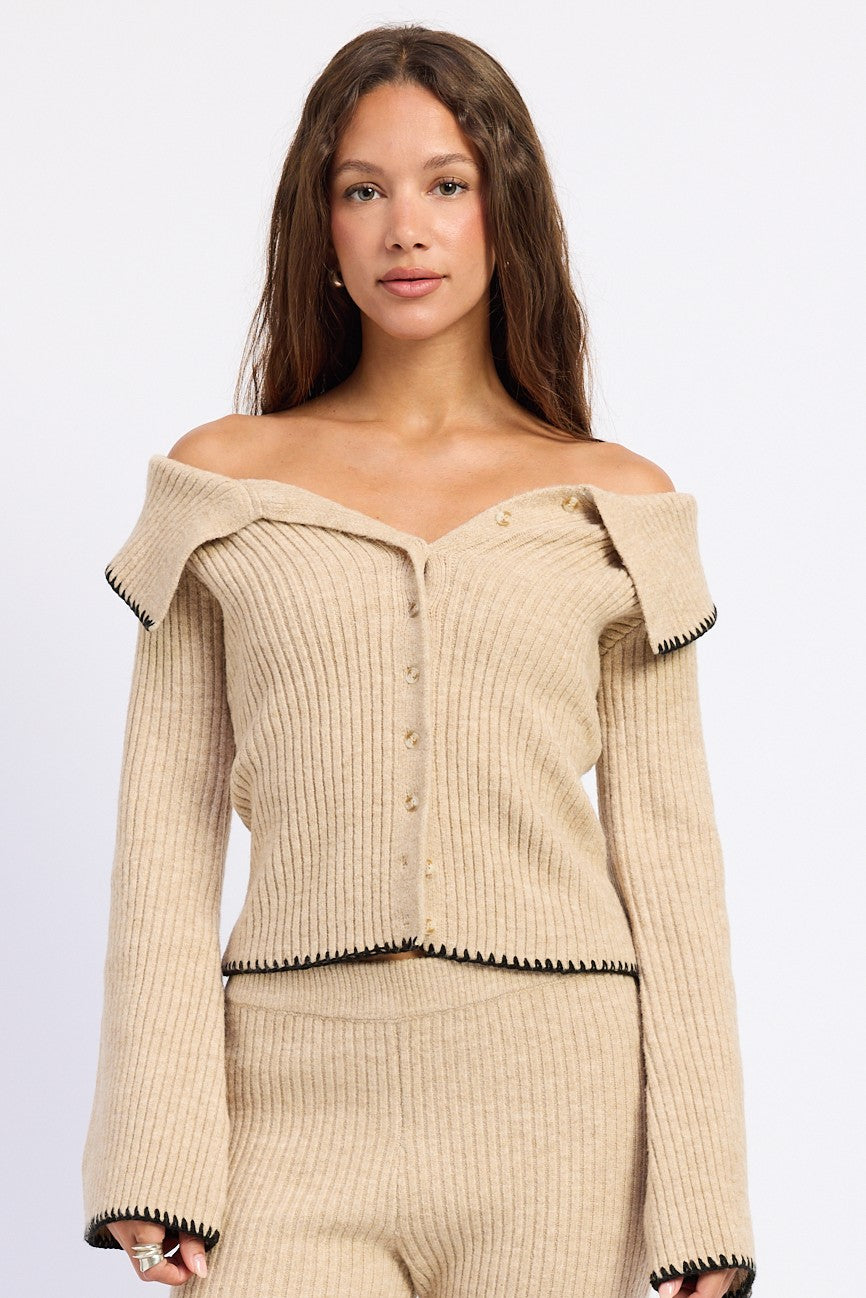 A woman modeling a beige ribbed knit off-the-shoulder cardigan with black trim and matching knit pants against a plain light gray studio background.