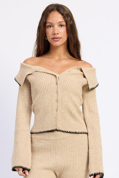 A woman modeling a beige ribbed knit off-the-shoulder cardigan with black trim and matching knit pants against a plain light gray studio background.