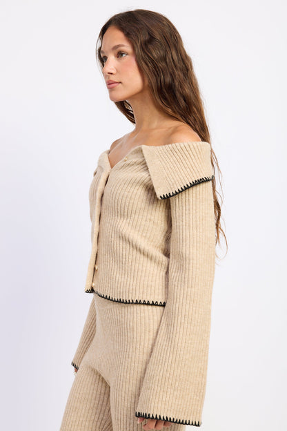 A woman wearing a beige ribbed knit off-the-shoulder cardigan and matching knit pants, shown in profile against a plain light background.