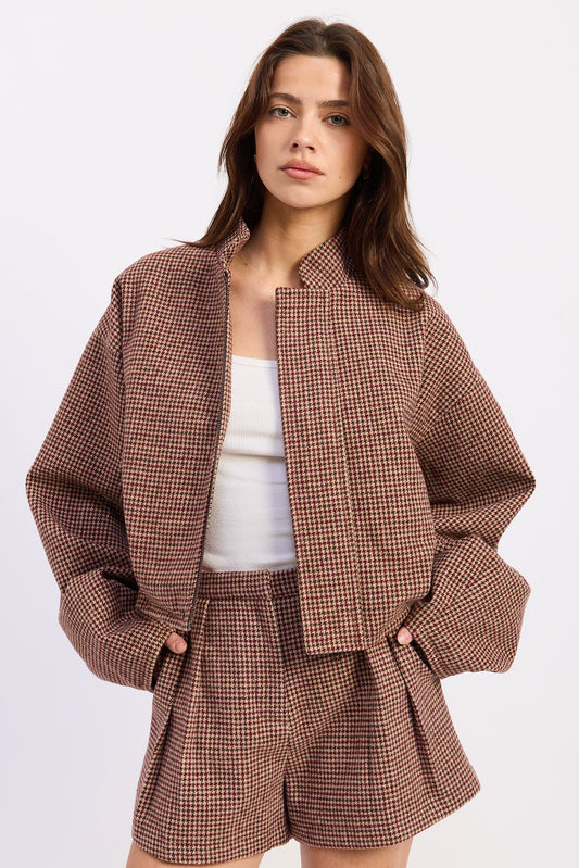 A woman modeling a brown and beige houndstooth cropped jacket and matching shorts over a white tank top, photographed against a clean white studio background.