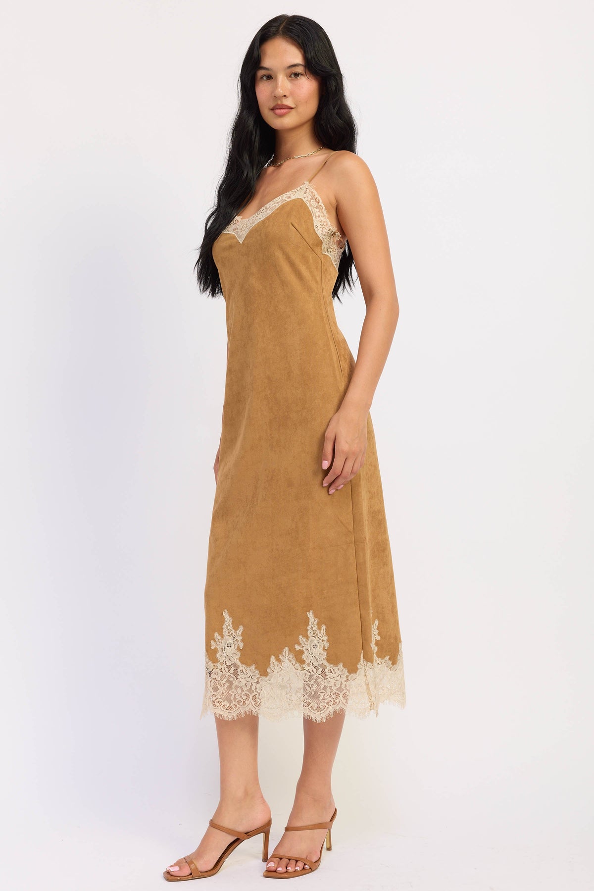 A woman modeling a tan slip dress with cream lace trim on the neckline and hem, paired with tan heeled sandals, against a plain white studio background.