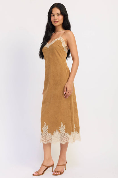 A woman modeling a tan slip dress with cream lace trim on the neckline and hem, paired with tan heeled sandals, against a plain white studio background.