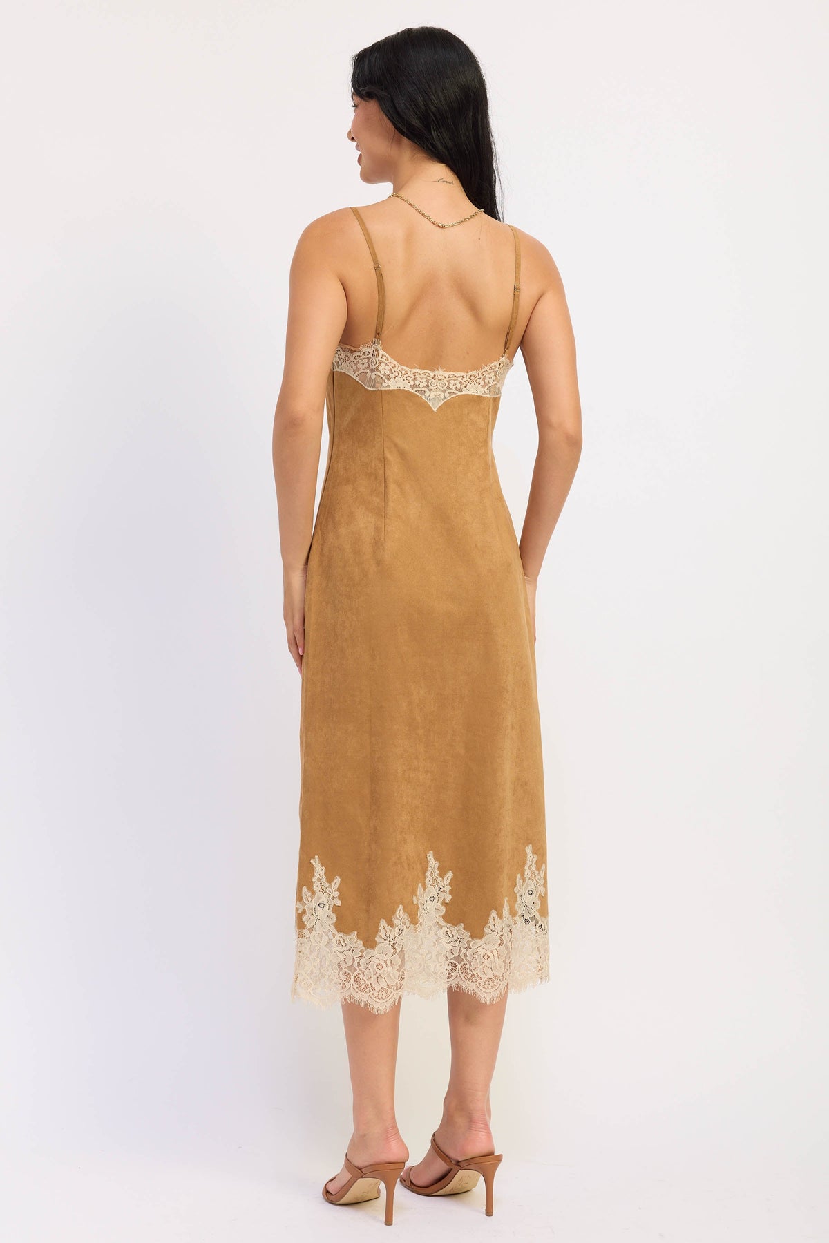 A woman shown from the back wearing a tan slip-style midi dress with cream lace trim at the neckline and hem, paired with tan heeled sandals against a plain light background.