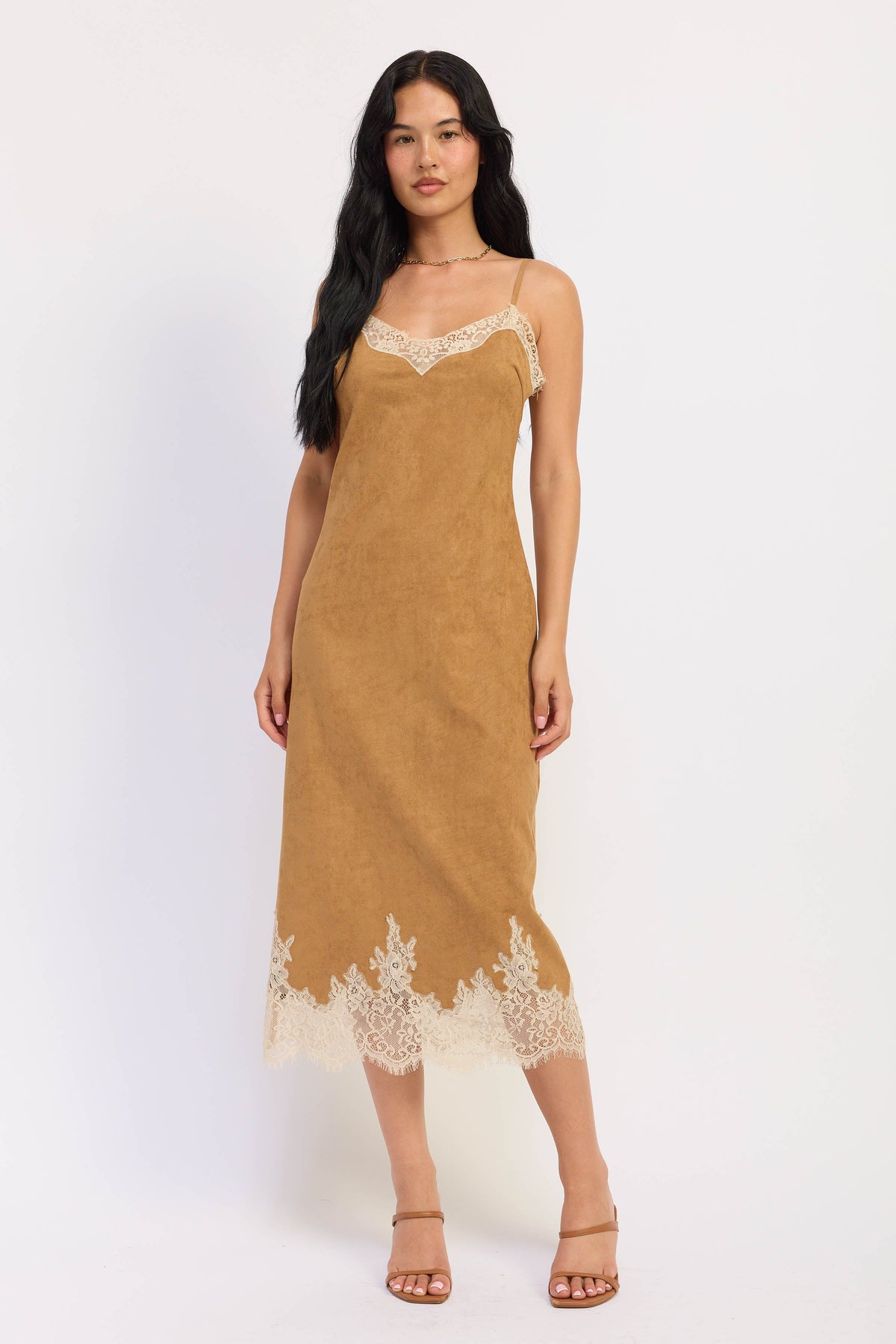 A woman modeling a tan slip-style midi dress with cream lace trim at the neckline and hem, standing against a plain light background.