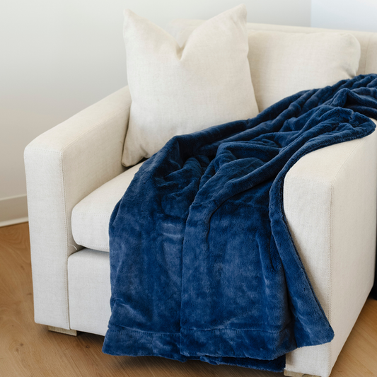 Blue LUSH THROW BLANKET draped over a beige armchair in a room with wooden flooring.