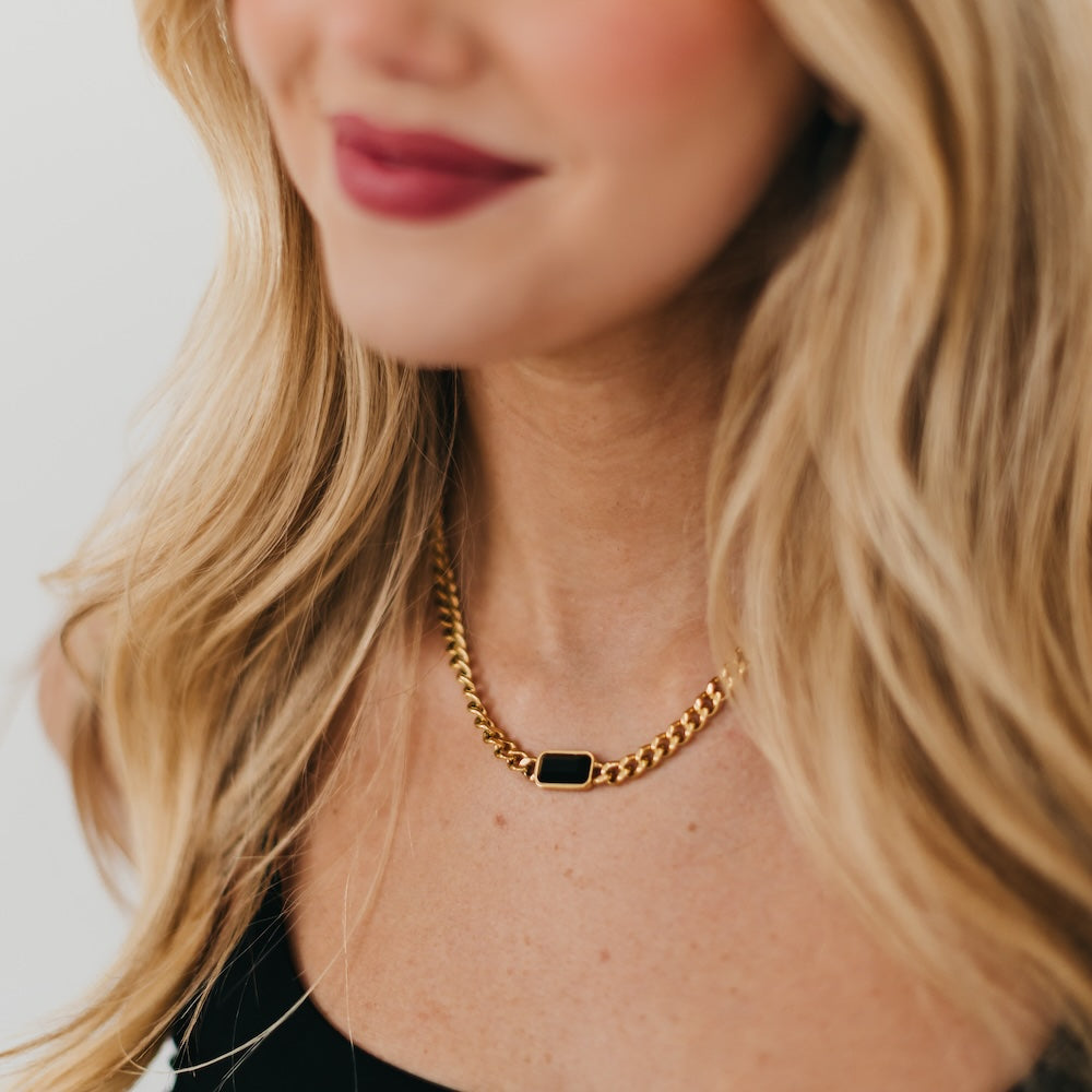 Close-up of a person wearing a gold chain necklace with a black pendant on a neutral background