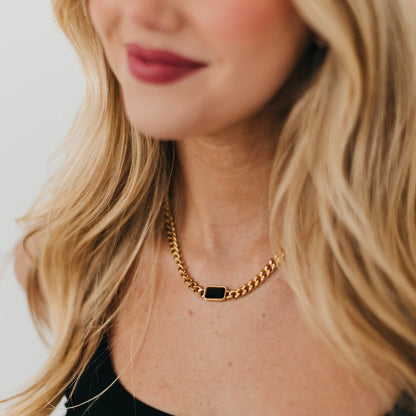 Close-up of a person wearing a gold chain necklace with a black pendant on a neutral background