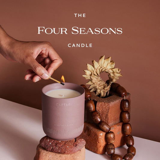 Hand lighting a candle labeled 'The Four Seasons Candle' with decorative elements on a brown background.