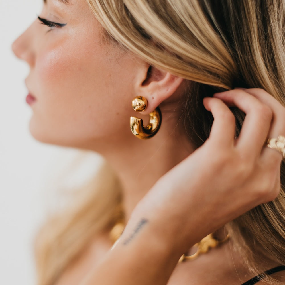 Close-up of a woman wearing gold hoop earrings and a ring, with a blurred background.