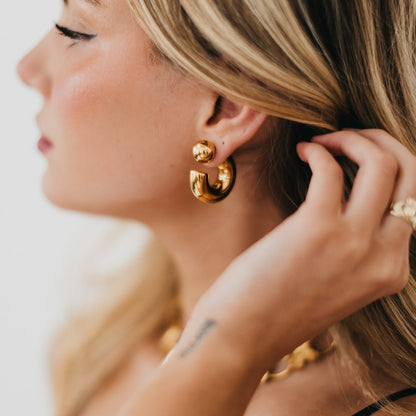 Close-up of a woman wearing gold hoop earrings and a ring, with a blurred background.
