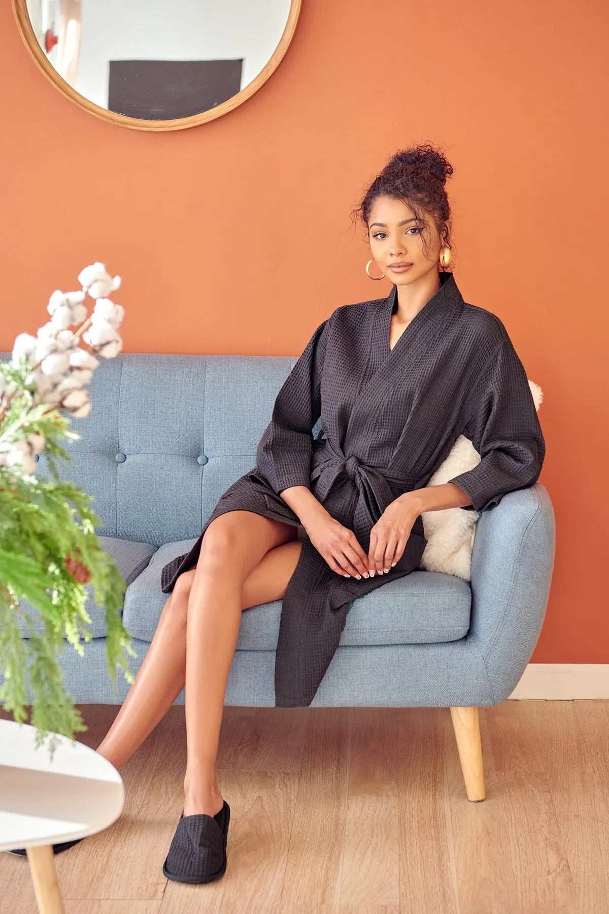 Woman in a dark Waffle Kimono Knee-Length robe  sitting on a light blue sofa against an orange wall.