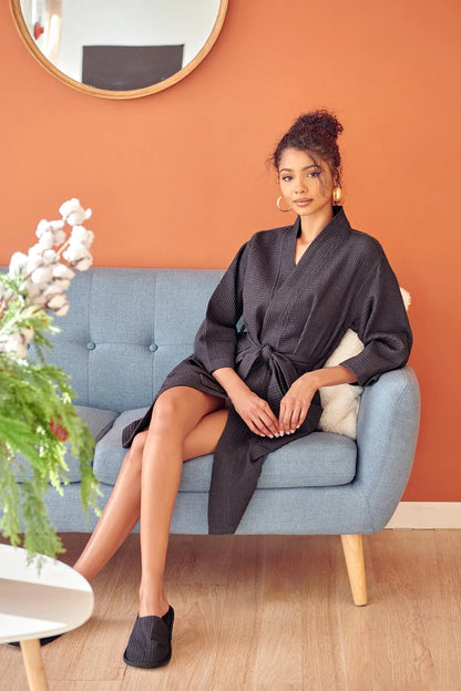 Woman in a dark Waffle Kimono Knee-Length robe  sitting on a light blue sofa against an orange wall.