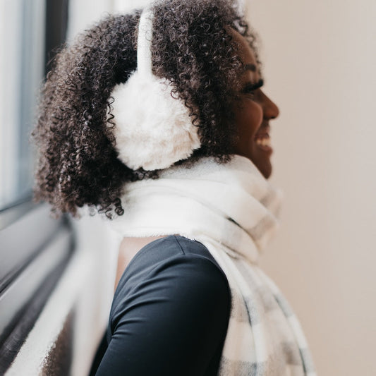 Woman wearing white ear muffs and a scarf, looking out a window.