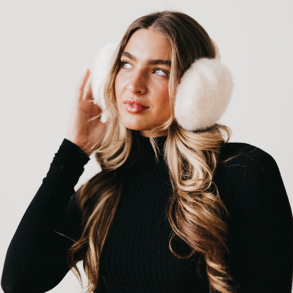 Woman wearing white fur ear muffs against a plain background