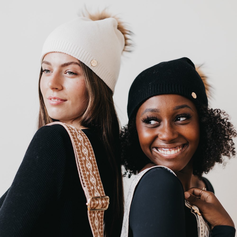 Two women wearing knit hats and smiling against a plain background